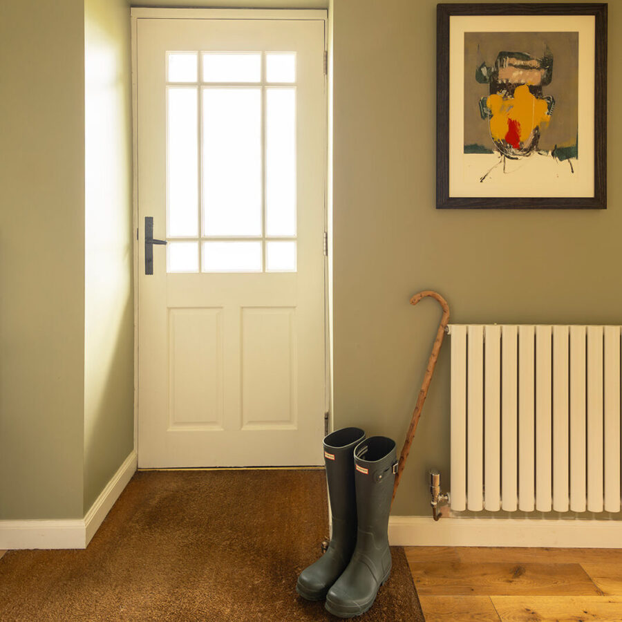 Inside the front door at the Croft House cottage. A glazed wooden door, painted white in a hall with soft sage green walls. Wellies and a can by the door.